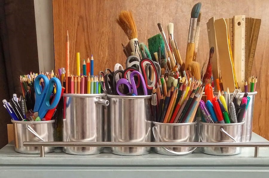 De nombreuses boîtes contenant des fournitures artistiques telles que des ciseaux, des pinceaux, des règles, des stylos et des crayons de couleur sont posées sur une étagère.