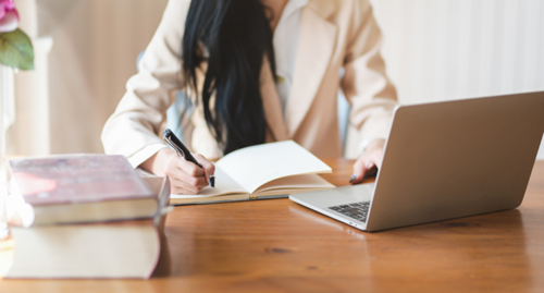 A person's mid-section visible behind a desk on which they are writing in a notebook. Also on the desk is an open laptop and two stacked books.
