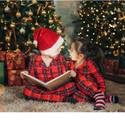 Two children wearing plaid pajamas and reading a book together while sitting in front of decorated christmas trees.