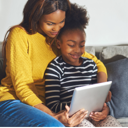A woman and a young child sit together on a couch reading a tablet. 