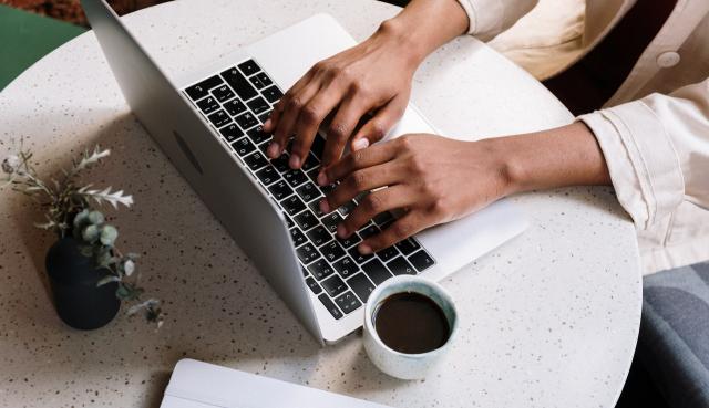 A Black woman typing on a laptop keyboard with a coffee and a plant resting next to the computer