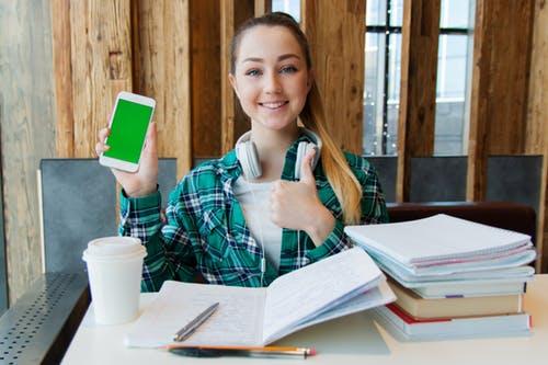 Teenage girl holding iphone sitting in front of books on a desk.